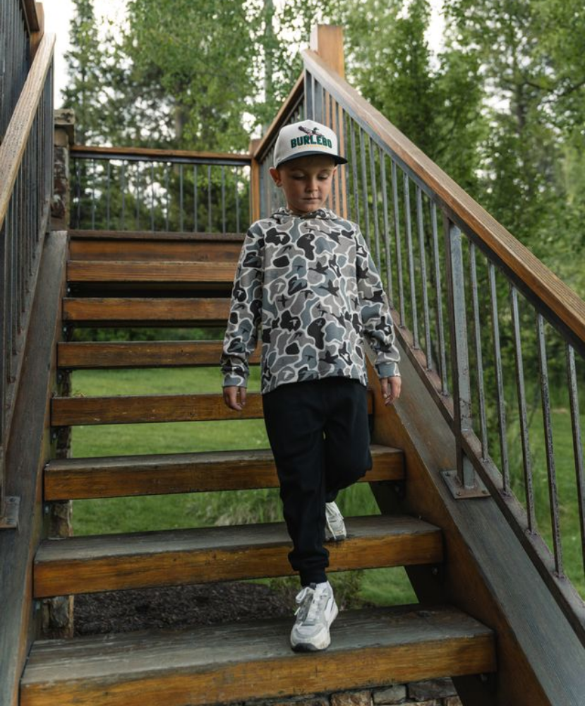 A child walking down outdoor wooden stairs wearing a gray and white camouflage hoodie, black pants, white sneakers, and a white baseball cap with a green logo.