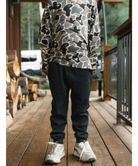A person standing on a wooden deck wearing a gray, black, and white camouflage long-sleeve shirt and dark jogger pants with white sneakers.