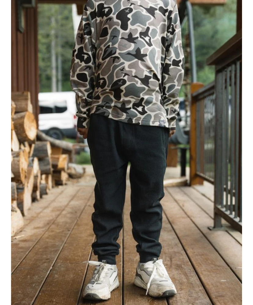 A person standing on a wooden deck wearing a gray, black, and white camouflage long-sleeve shirt and dark jogger pants with white sneakers.