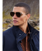 Man wearing Krewe sunglasses and a dark jacket in a natural setting
