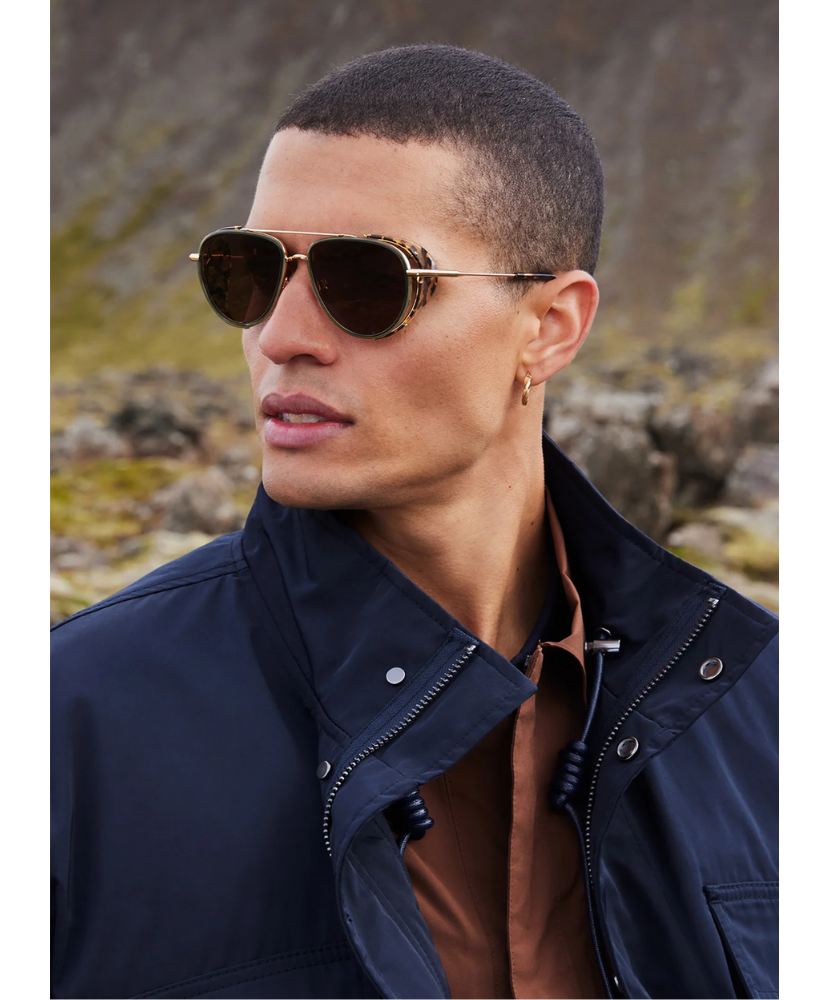 Man wearing Krewe sunglasses and a dark jacket in a natural setting