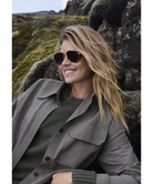 Woman wearing Krewe sunglasses and a coat in a natural setting with rocks and grass.