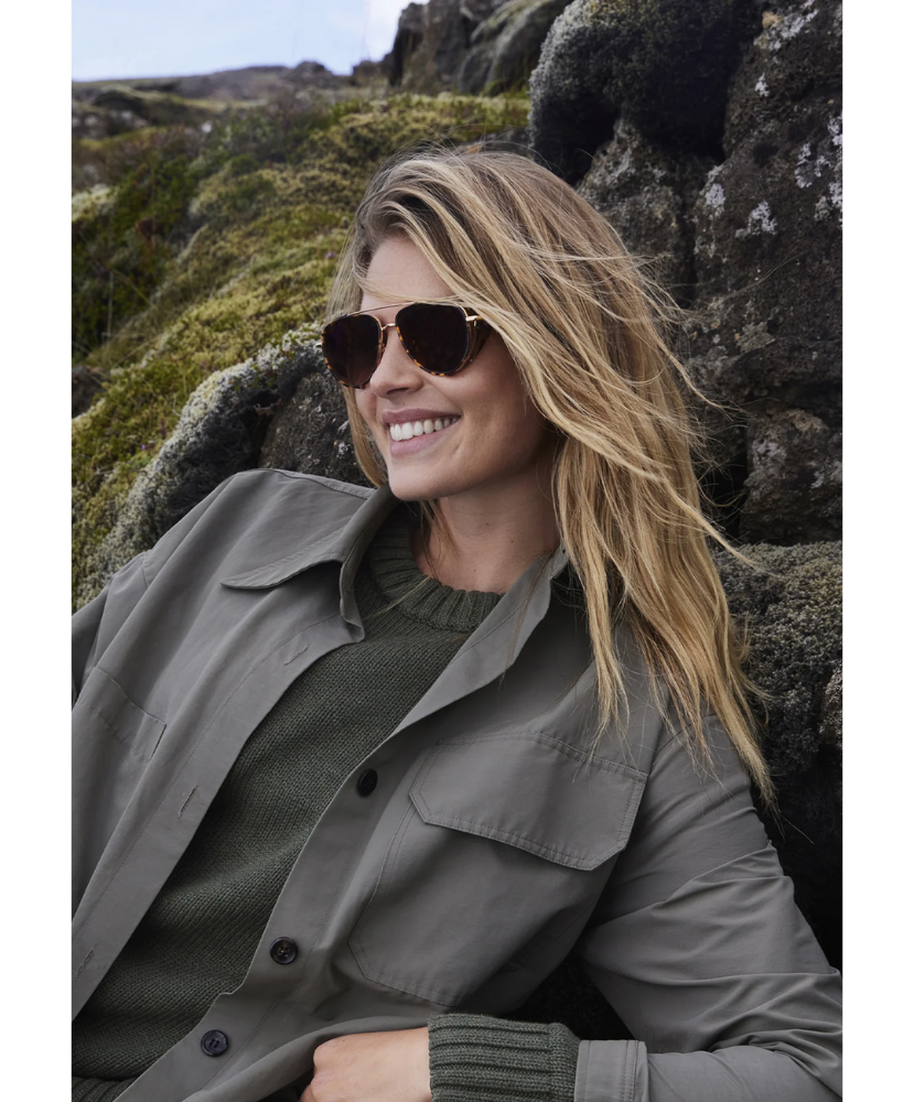 Woman wearing Krewe sunglasses and a coat in a natural setting with rocks and grass.