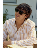 Man wearing Krewe sunglasses and a striped shirt sitting outdoors.