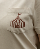 Beige t-shirt with a red and white striped tent design on the chest pocket.