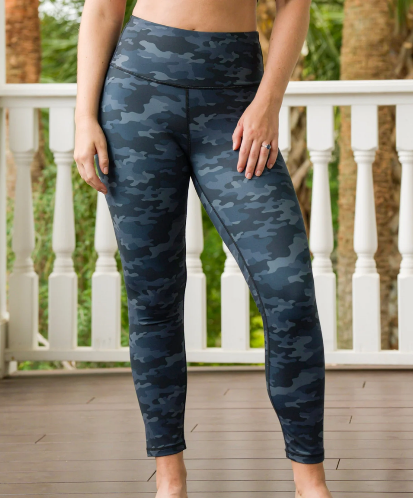 A person wearing high-waisted leggings with a dark blue and gray camouflage pattern, standing on a wooden deck with a white railing and green foliage in the background.