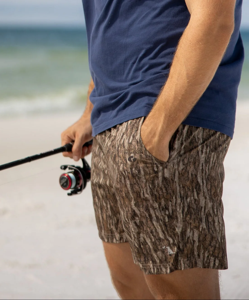 A man on a beach wearing brown and beige camouflage fishing shorts, a navy blue t-shirt, and holding a fishing rod.