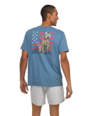 Man wearing a blue t-shirt with a graphic of a dog and American flag on the back, standing against a white background.