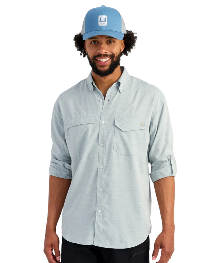 Man wearing a light blue HUK button-up shirt and blue cap on a white background