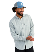 Man wearing a light blue HUK button-up shirt and blue cap on a white background