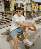 Man sitting on a wooden bench outdoors, wearing a henley holding a drink.