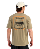 Man wearing a beige t-shirt with Huk & Co fishing supplies logo on the back. Bass graphic.