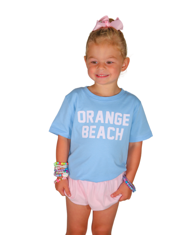 Young girl wearing a light blue 'Orange Beach' t-shirt and pink shorts on a white background