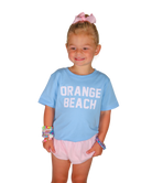 Young girl wearing a light blue 'Orange Beach' t-shirt and pink shorts on a white background