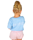 Child wearing a light blue shirt with 'SHADES SUNGLASSES' text and pink shorts on a white background