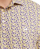 Close-up of a men's short-sleeve button-up shirt with a yellow base and an all-over purple and gold pattern of shopping bags and gift boxes, featuring a white embroidered Lacoste crocodile logo on the chest.