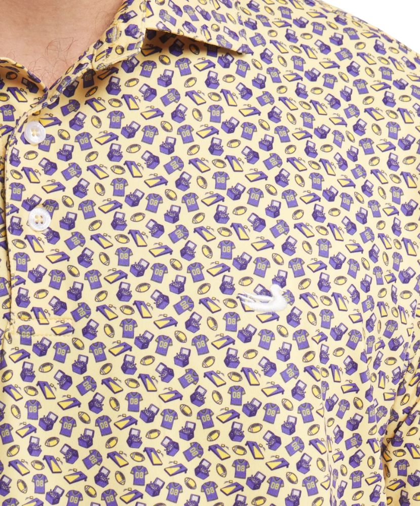 Close-up of a men's short-sleeve button-up shirt with a yellow base and an all-over purple and gold pattern of shopping bags and gift boxes, featuring a white embroidered Lacoste crocodile logo on the chest.