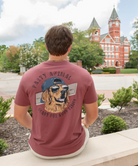 Person wearing a maroon t-shirt with a dog graphic and text, sitting in front of a red brick building.