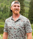 Man wearing a camouflage polo and cap outdoors with greenery in the background