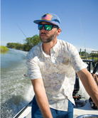Man on a boat wearing a cap with an American flag, sunglasses, and a patterned shirt.