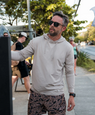 Man wearing a BEIGE hoodie and camouflage shorts outdoors with trees and people in the background.