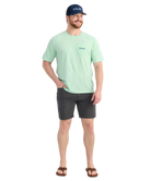 Man wearing a light green HUK t-shirt and dark shorts with a white background