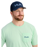 Man wearing a green Huk shirt and navy cap on a white background