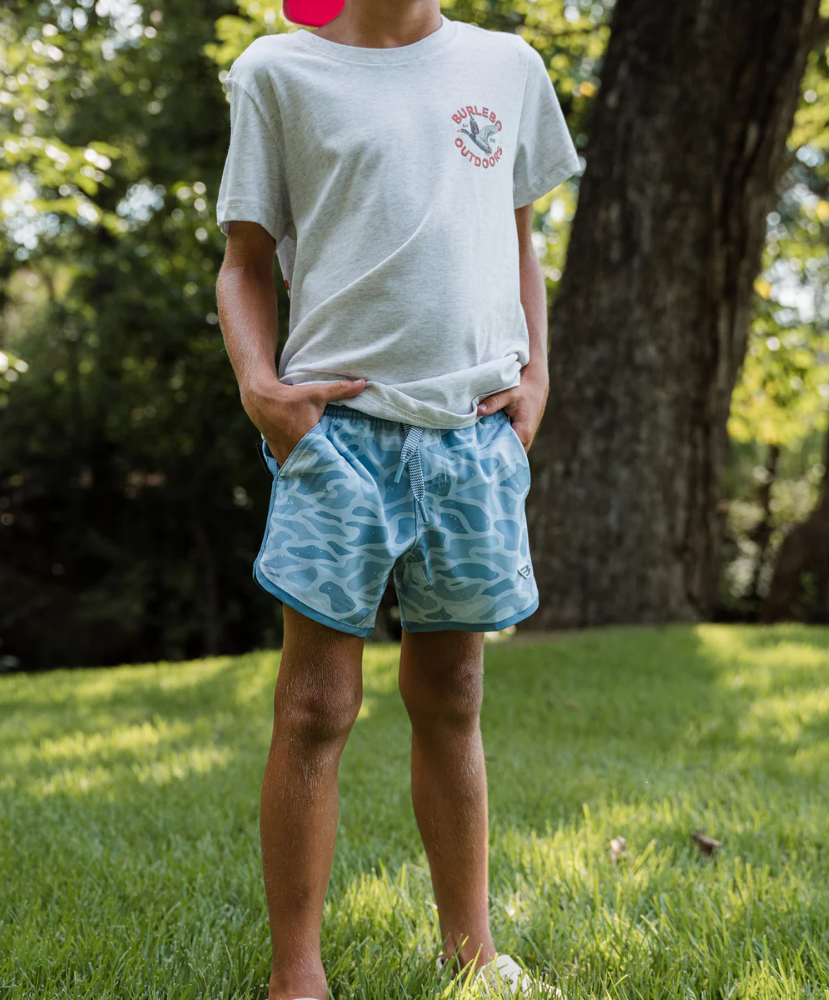 Boy wearing a light gray t-shirt and blue camo swim trunks standing outdoors with trees in the background.