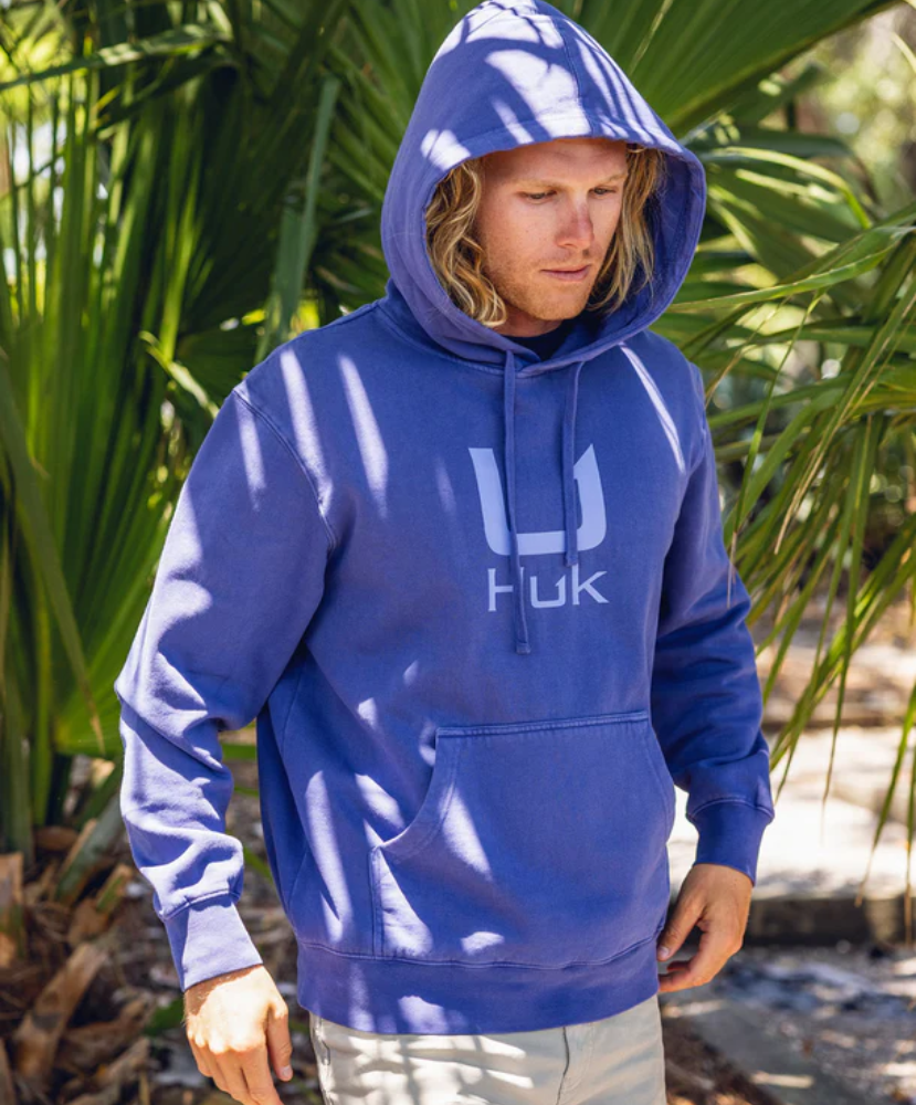 Person wearing a blue hoodie with a HUK logo, standing outdoors among green plants.