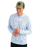 Man wearing a light blue camouflage hoodie with 'Huk' logo and a green cap on a white background