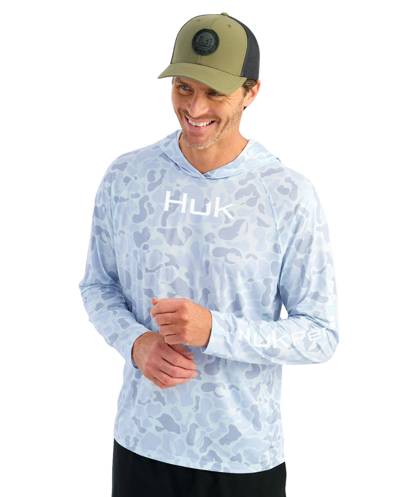 Man wearing a light blue camouflage hoodie with 'Huk' logo and a green cap on a white background