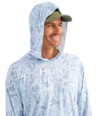Man wearing a light blue camouflage hoodie with 'Huk' logo and a green cap on a white background