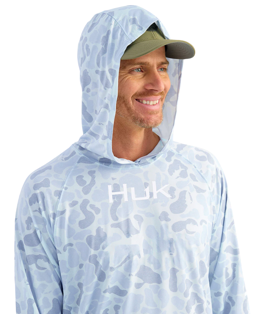 Man wearing a light blue camouflage hoodie with 'Huk' logo and a green cap on a white background