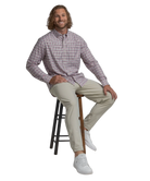 Man sitting on a stool wearing a checkered shirt and beige pants against a white background