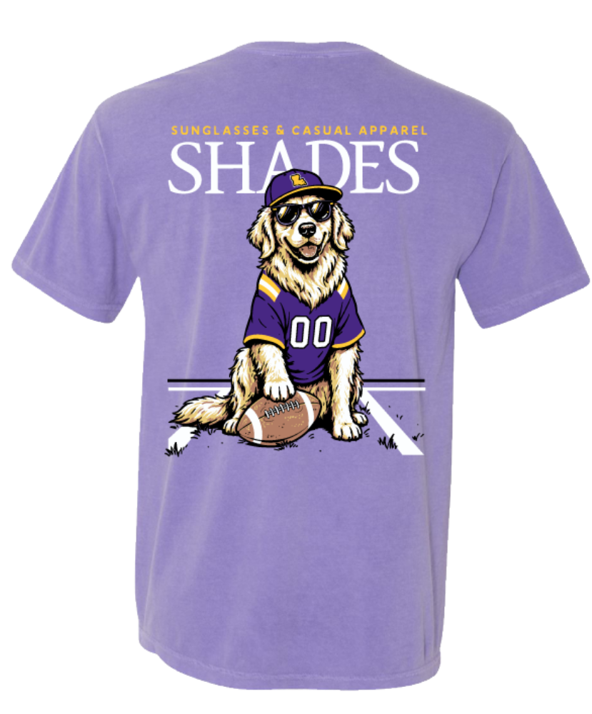 A purple short-sleeve t-shirt with a large graphic on the back. The graphic features the text 'SUNGLASSES & GUNGLASSES APPAREL SHADES' in white and yellow, with a cartoon golden retriever wearing a purple and gold football jersey and a matching cap, sitting on a football.
