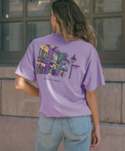 Purple Mardi Gras t-shirt with a colorful graphic of a New Orleans street scene on the back, on a white background.
