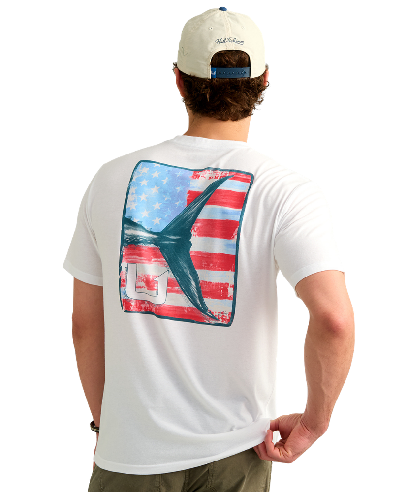 Man wearing a USA HUK white t-shirt with an American flag and marlin tail graphic design on the back, standing against a white background
