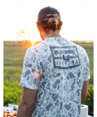 Man wearing a camouflage t-shirt with 'Nuk Performance Fishing' logo, standing outdoors with sunset in the background.