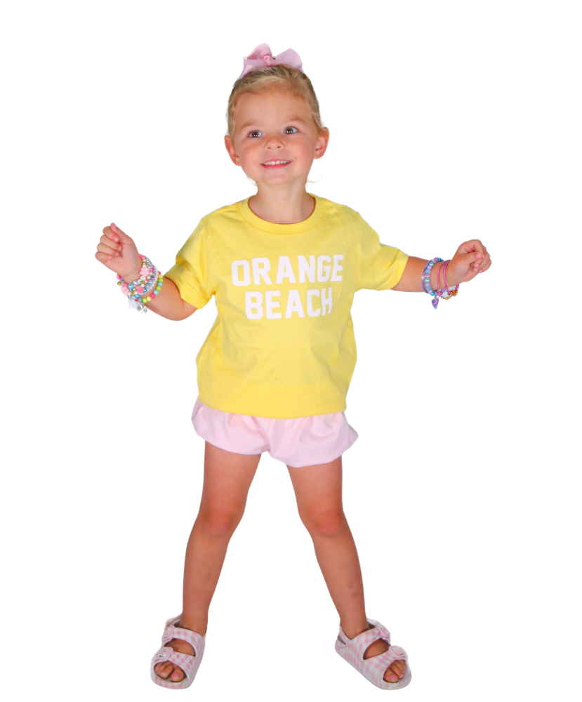 Young girl wearing a yellow 'Orange Beach' shirt with pink shorts on a white background