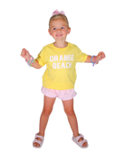 Young girl wearing a yellow 'Orange Beach' shirt with pink shorts on a white background
