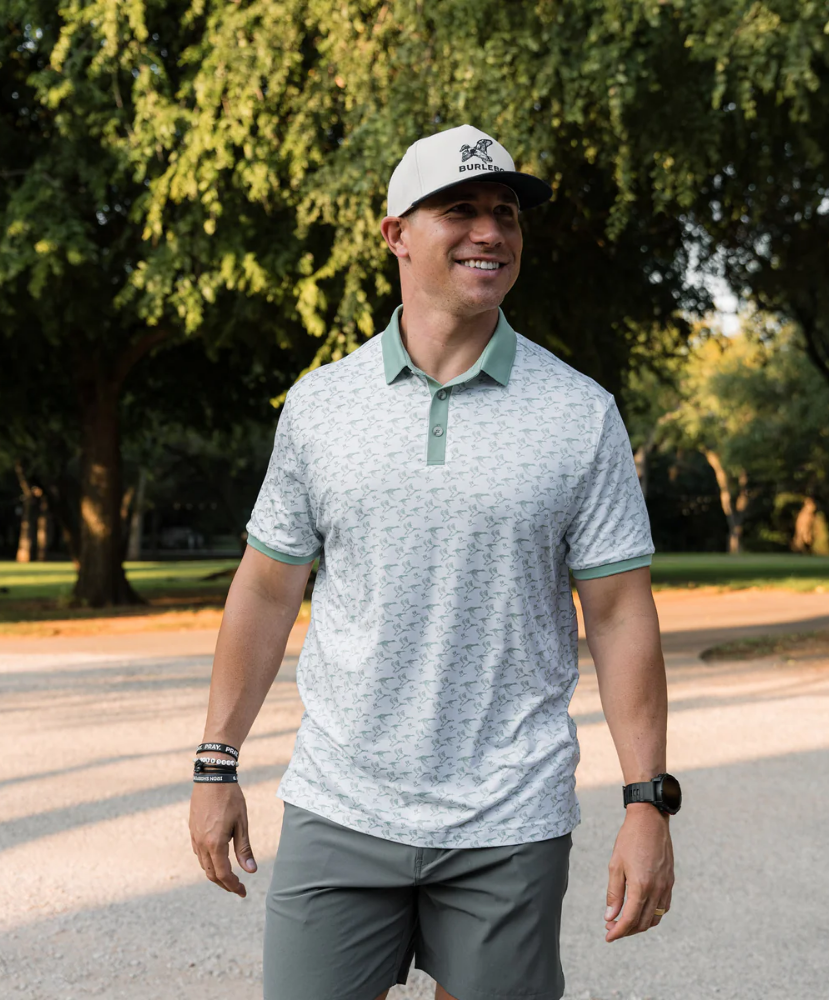 Man wearing a white duck and waterfowl patterned polo shirt with green accents on a white background